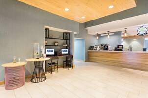 Un interior de cafetería moderna con un mostrador de recepción de madera, zona de estar con mesas y sillas, y una barra con máquinas de café.