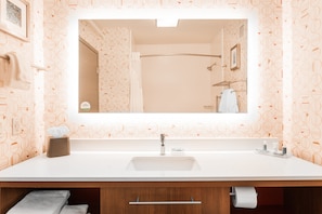 A bathroom with a large mirror, a sink, and a shower. There are towels and toiletries on the counter.