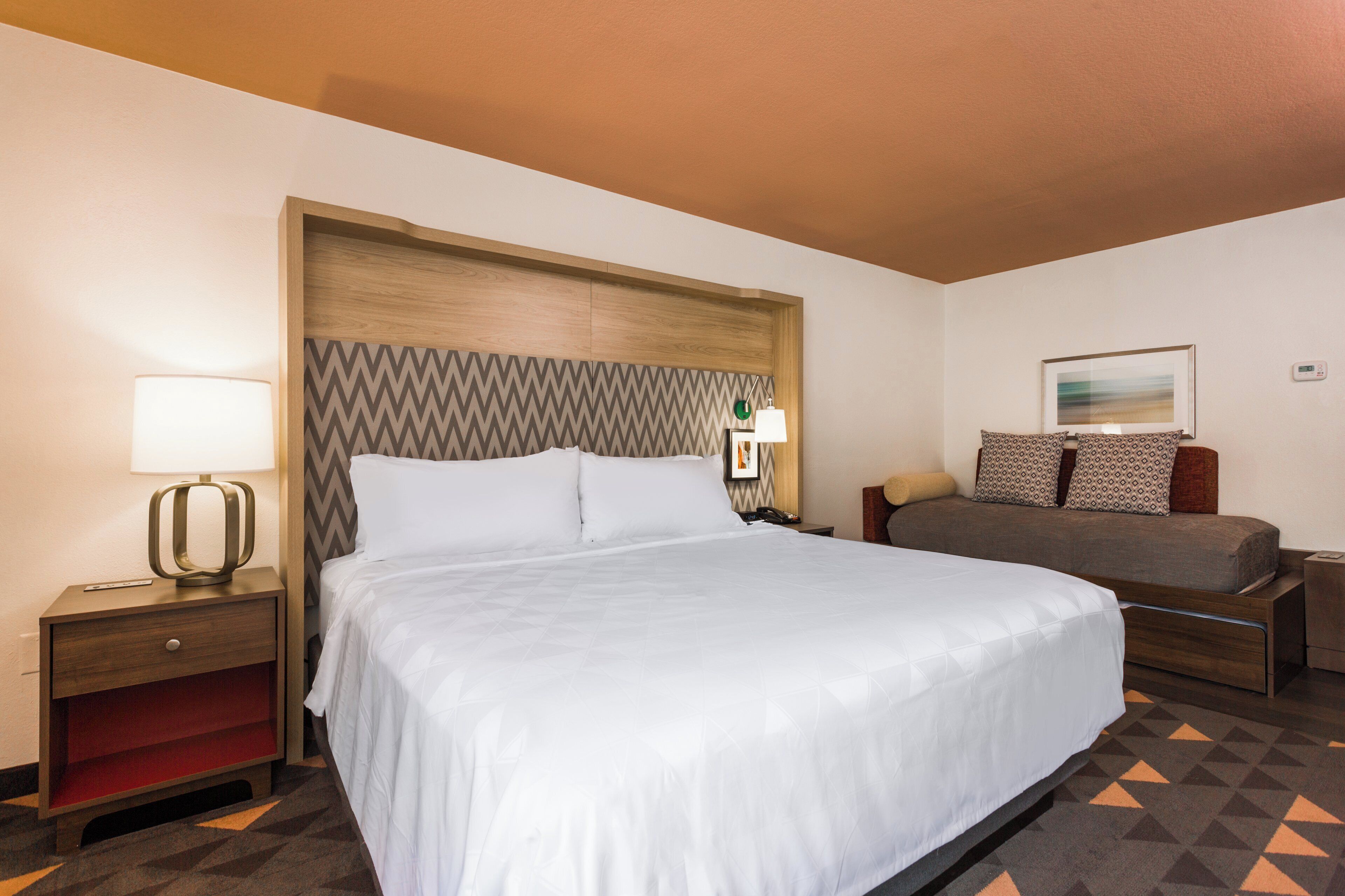 A hotel room with a large bed, a bedside table with a lamp, a small desk, and a chair.