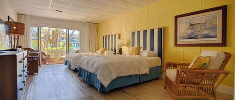 A hotel room with two beds, a wicker chair, a desk, and a large window with a view of palm trees.