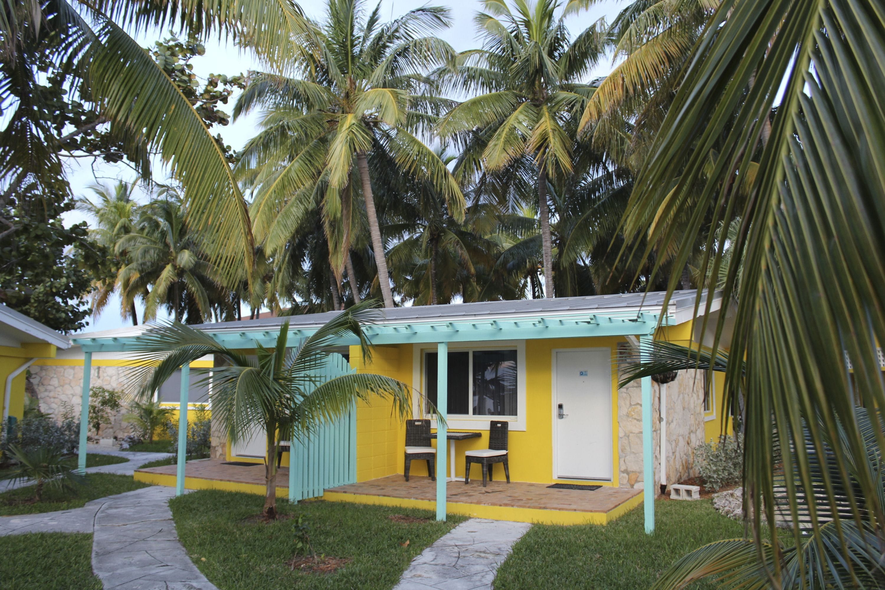 A small, yellow building with a covered patio area, surrounded by palm trees.