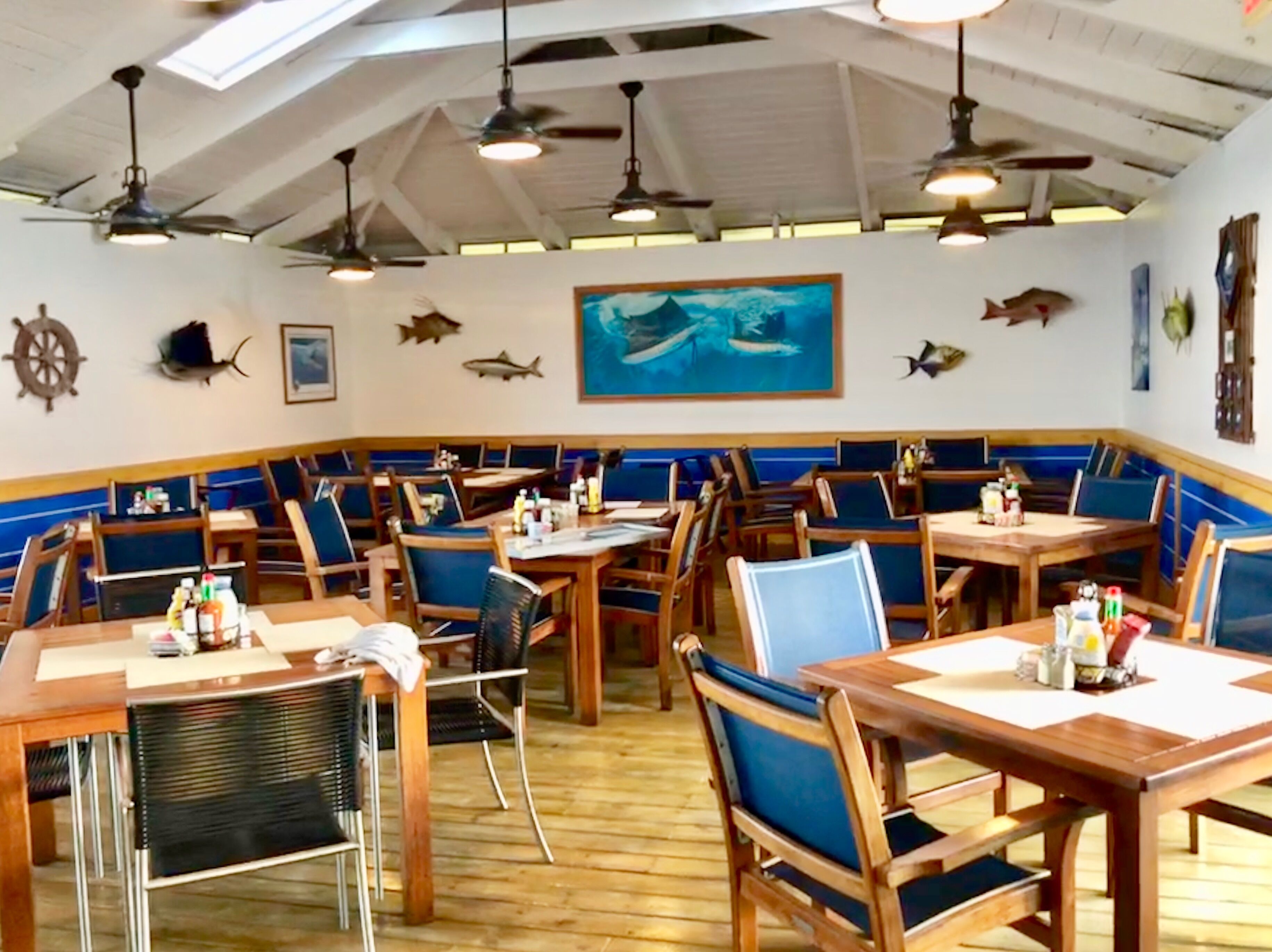 A restaurant with wooden tables and blue cushioned chairs, nautical-themed wall decorations, and ceiling fans.