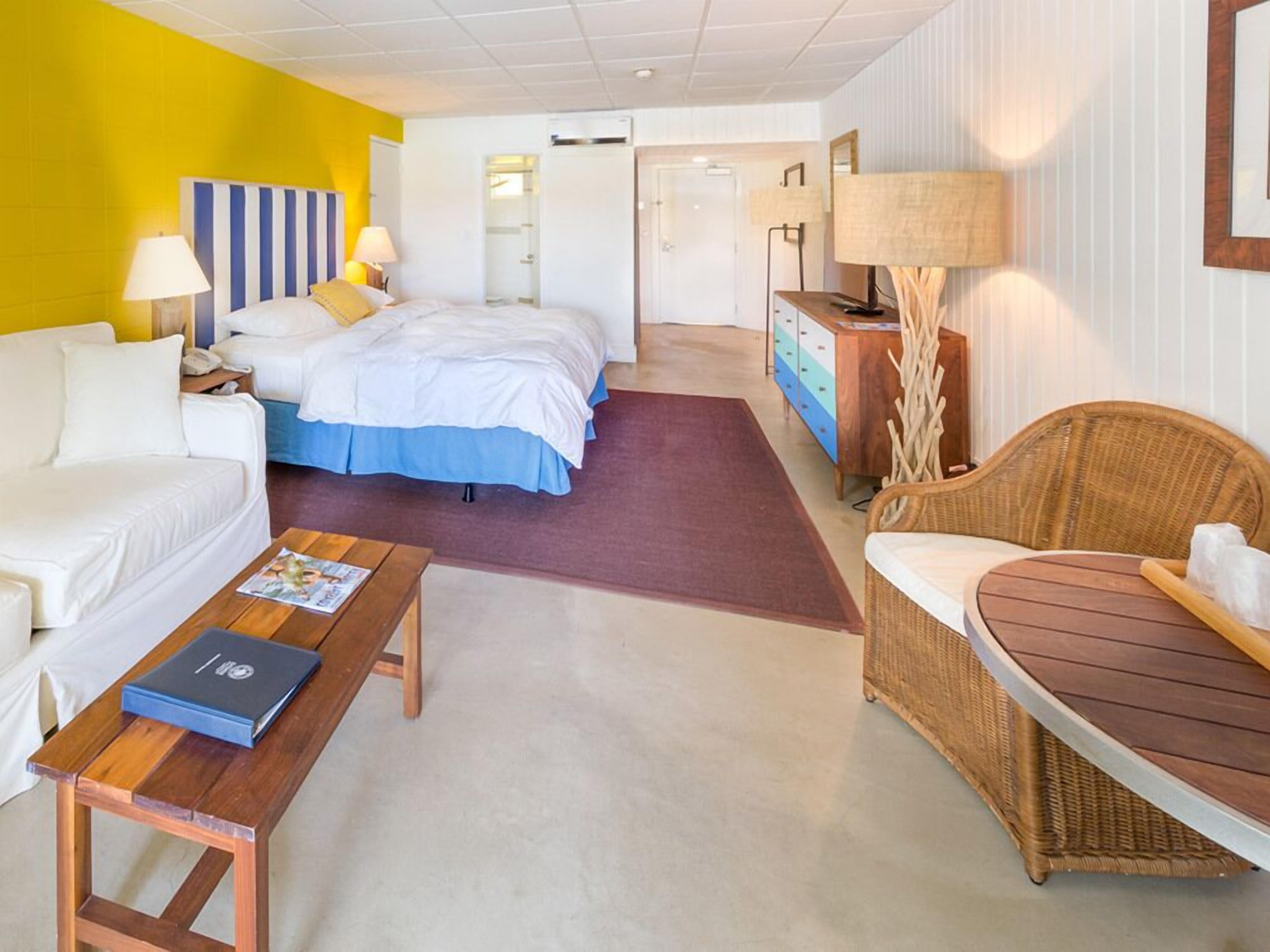 A hotel room with a bed, a sofa, a wooden table, a wicker chair, and a dresser.