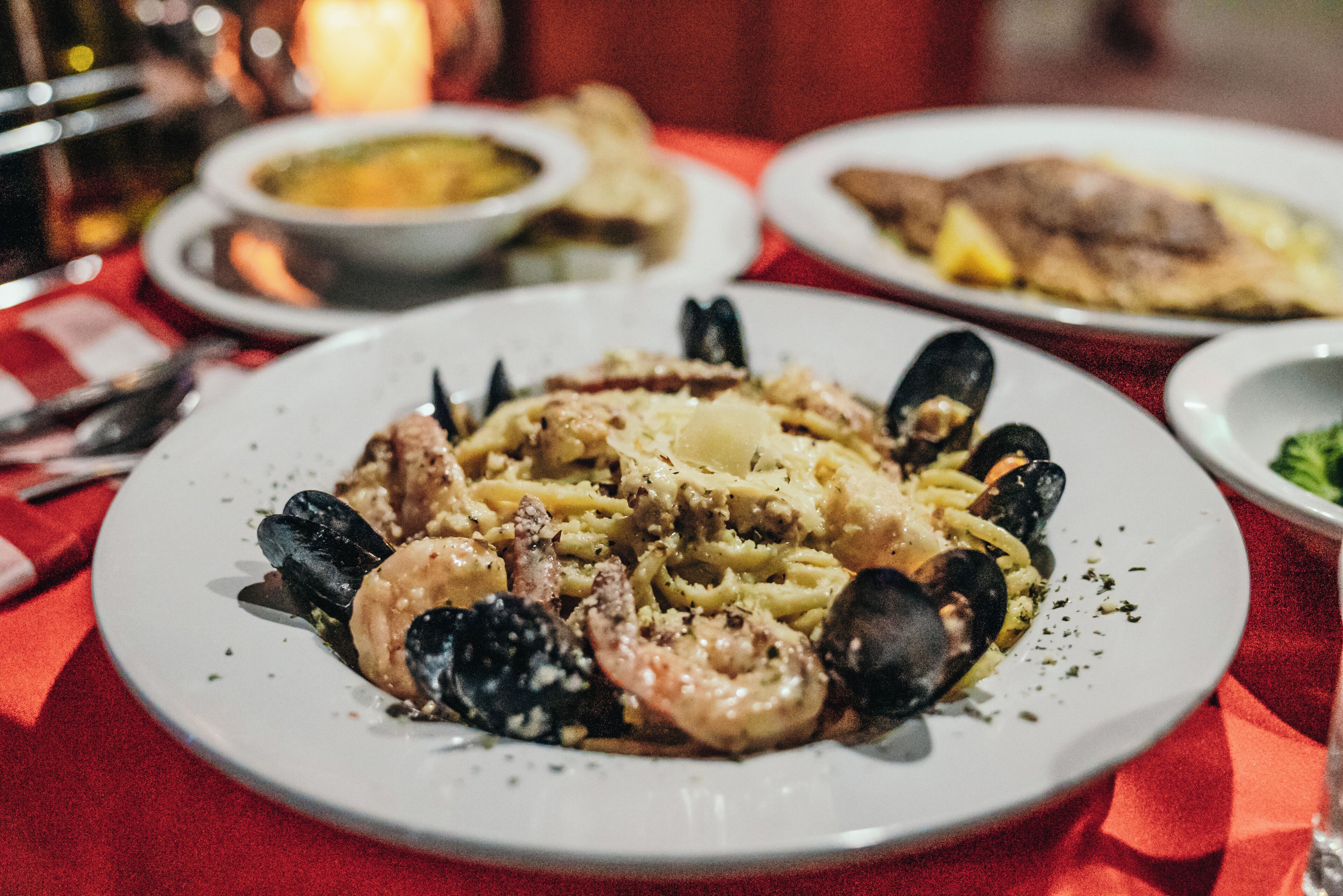 Une assiette de pâtes aux fruits de mer, avec des moules et des crevettes, servie dans une assiette blanche sur une table recouverte d’une nappe rouge.
