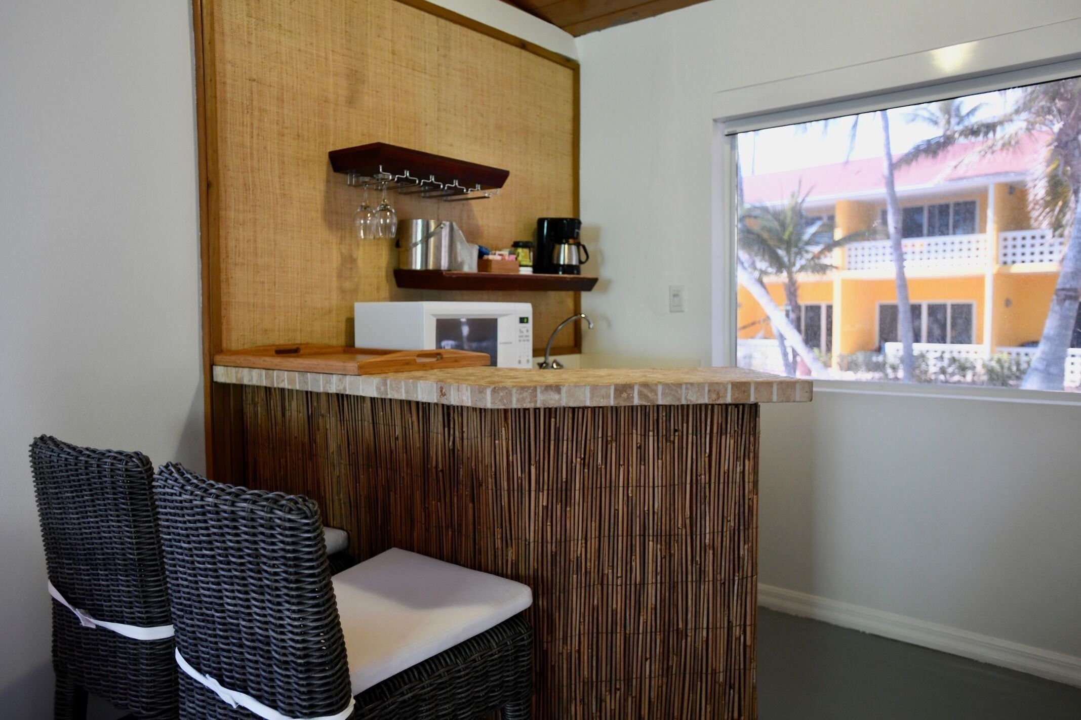 A kitchenette with a microwave, coffee maker, and wine glasses on a shelf. There is a wicker chair with a cushion and a window with a view of a yellow building.