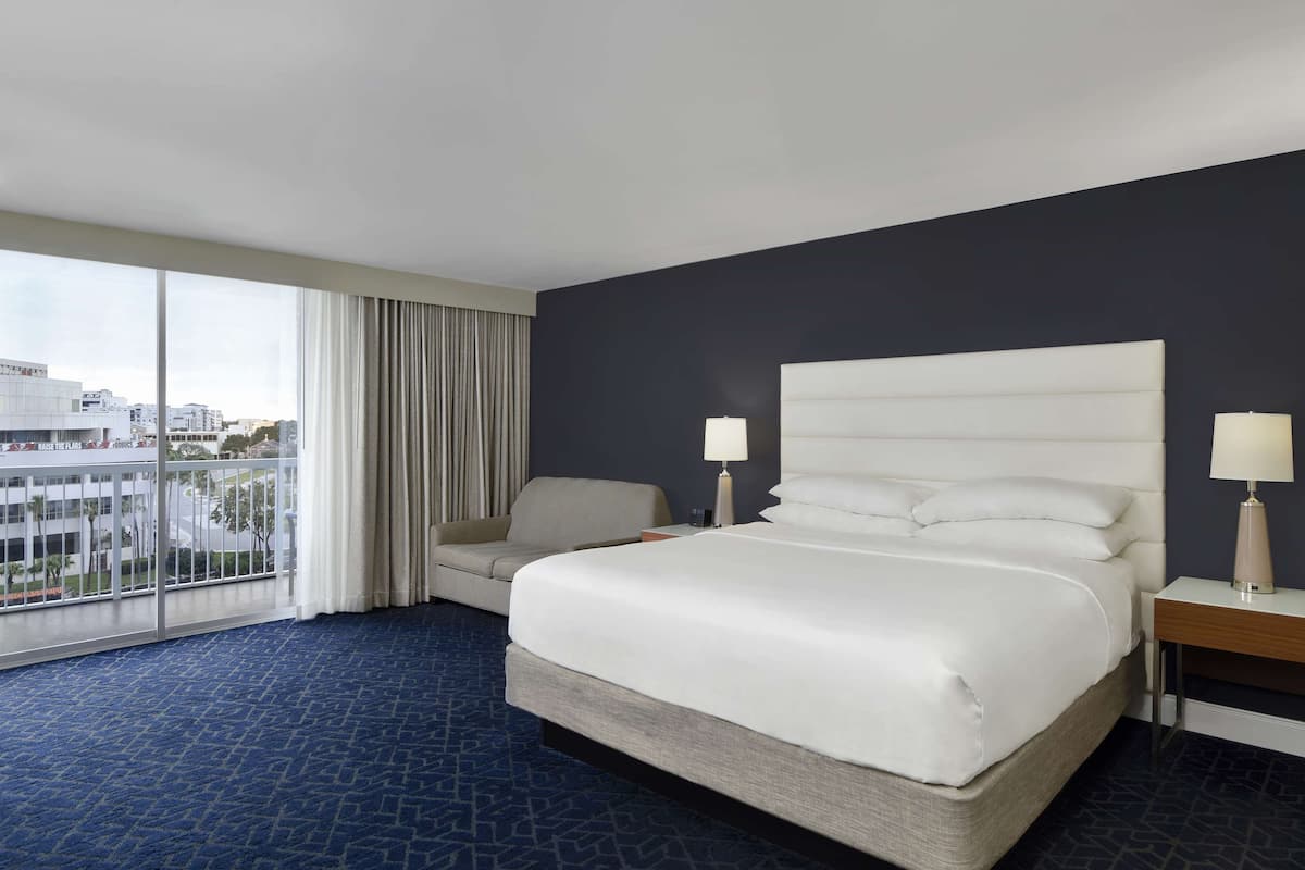 Une chambre d’hôtel avec un grand lit, un canapé, deux tables de chevet avec des lampes, une vue sur la ville grâce à de grandes fenêtres et un mur de couleur sombre.