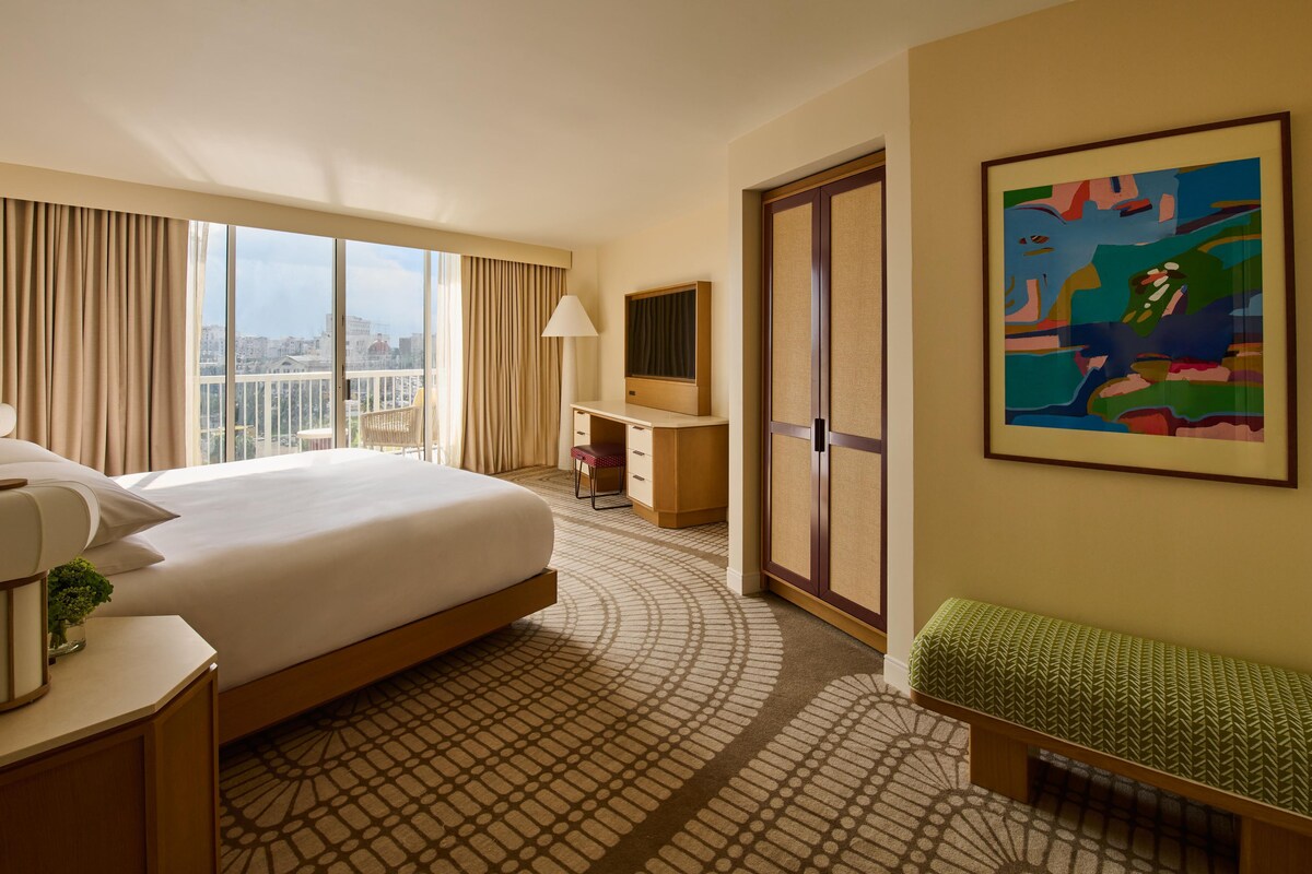 Habitación de hotel con una cama grande, un escritorio, una silla, una mesita con lámpara, un banco, un cuadro y vistas de la ciudad a través de la ventana.