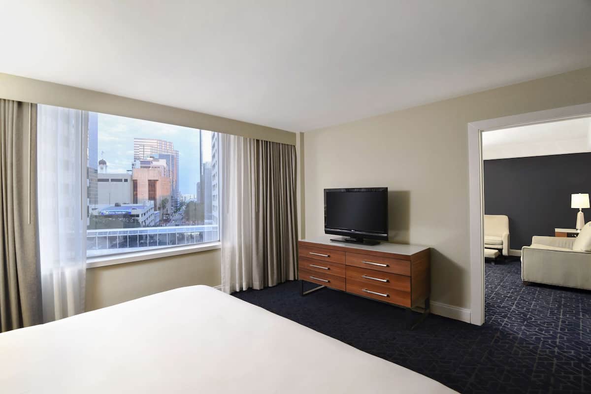 Une chambre d’hôtel avec un grand lit, un téléviseur posé sur une commode en bois, une vue sur la ville par une grande fenêtre et une porte donnant sur une autre pièce.