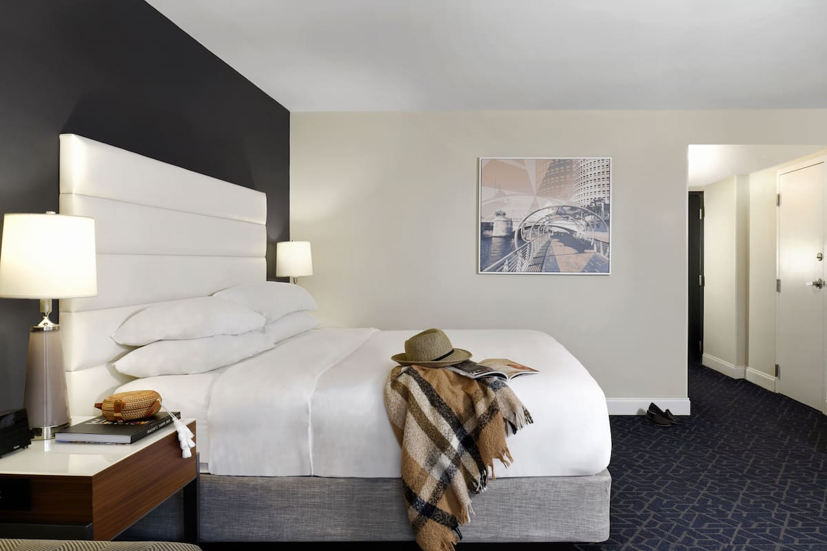 Une chambre d’hôtel avec un grand lit, une table de chevet avec une lampe, un magazine et un chapeau posé sur le lit.