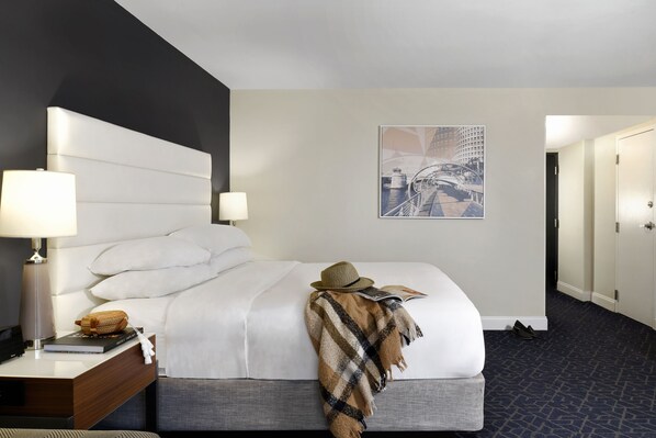 Habitación de hotel con una cama grande, una mesita de noche con una lámpara, una revista y un sombrero sobre la cama.