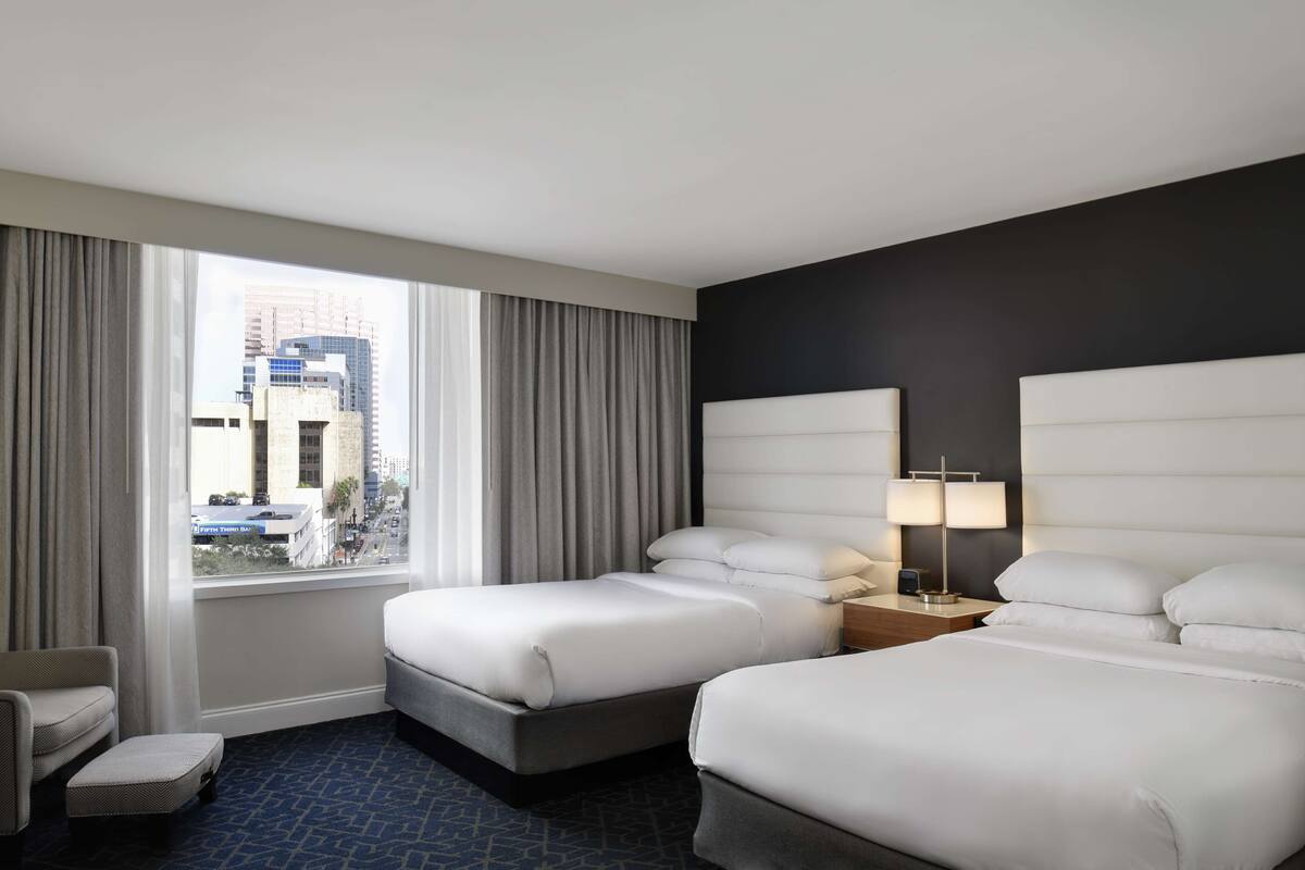 Une chambre d’hôtel avec deux lits, une chaise, une petite table et une vue sur le paysage urbain grâce à de grandes fenêtres.