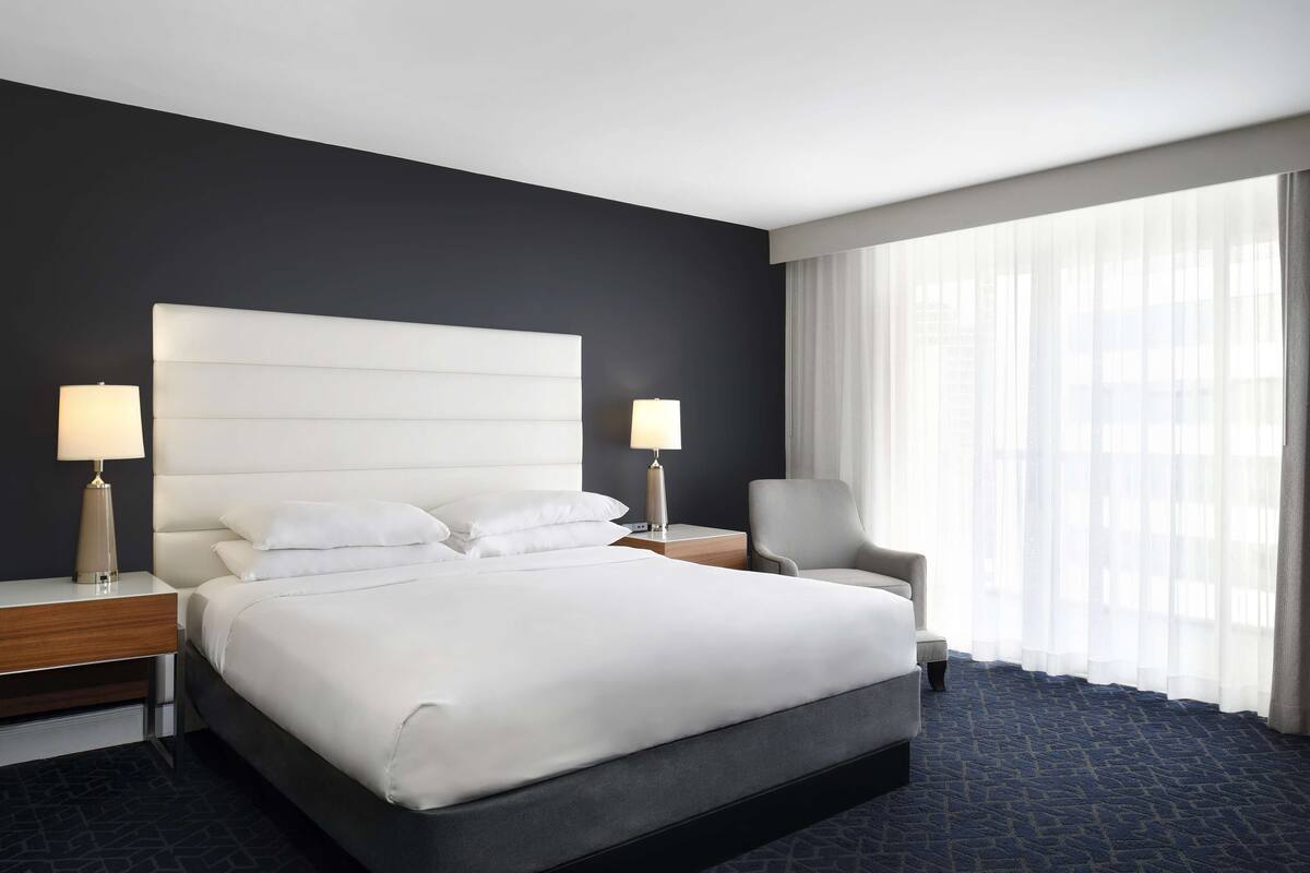 Une chambre d’hôtel avec un grand lit, deux tables de chevet avec des lampes, une chaise et une grande fenêtre avec des rideaux légers.