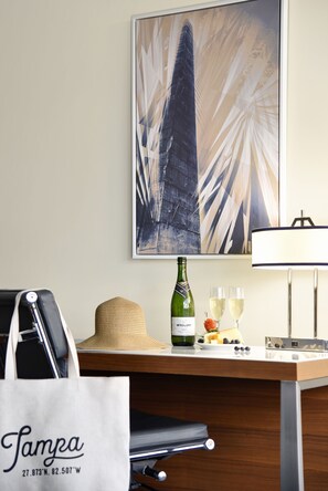 Hotelkamer met een groot abstract schilderij, een champagnefles, glazen, een strohoed en een witte tas met "Tampa" erop.