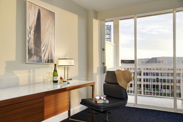 Una habitación de hotel con un escritorio, una silla, un ventanal con vistas a edificios y un cuadro en la pared.