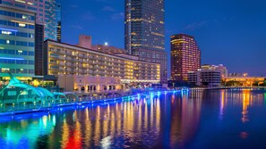 Een hotel aan het water, met meerdere verdiepingen, een zwembad met verlichte randen en een stadsachtergrond.