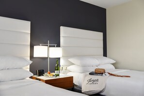 Una habitación de hotel con una cama grande, dos mesitas de noche, una lámpara, una botella de vino, copas y un bolso con el nombre 