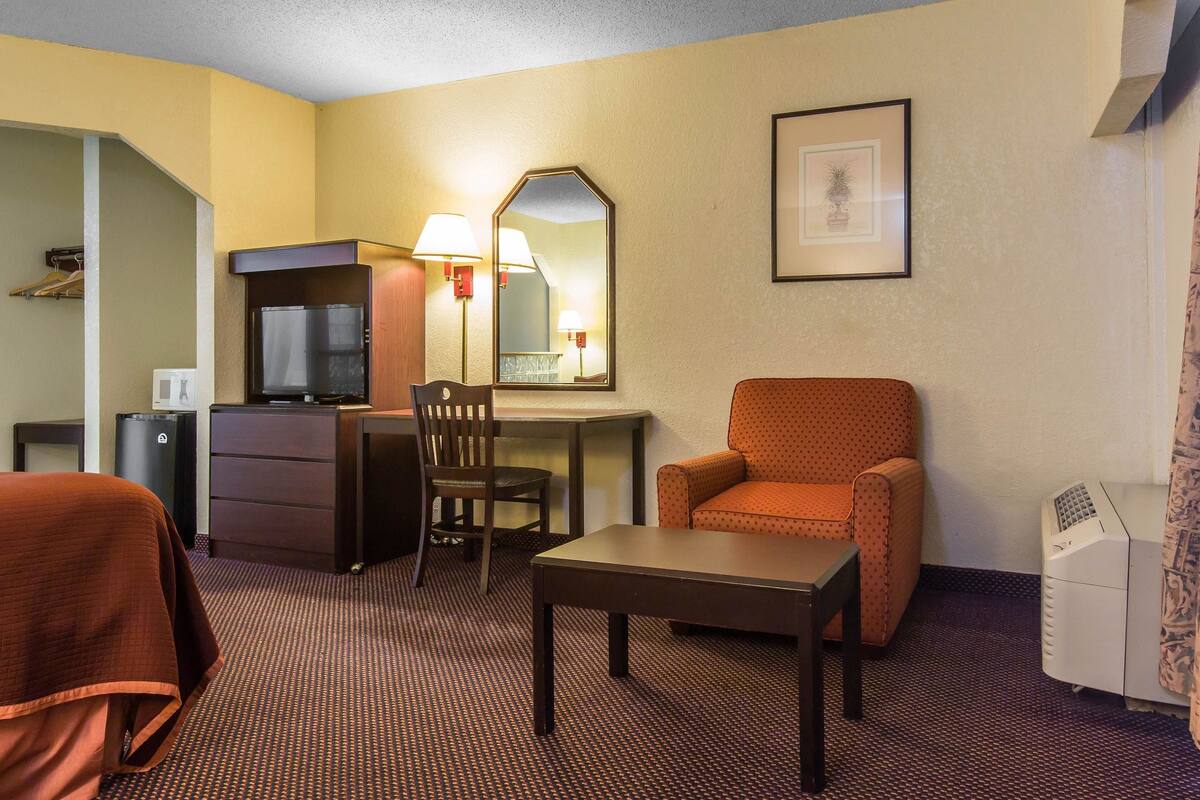 A hotel room with a bed, a desk with a chair, a television, a mirror, a framed picture on the wall, and a printer.