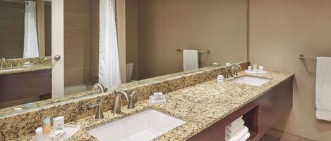 A hotel bathroom with a granite countertop, two sinks, a large mirror, and a shower area.