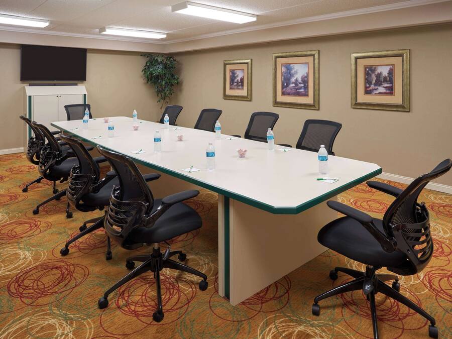 A conference room with a long table, six chairs, water bottles, a projector screen, framed pictures, and a carpeted floor.