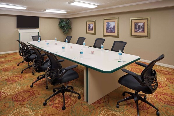 A conference room with a long table, six chairs, water bottles, a projector screen, framed pictures, and a carpeted floor.