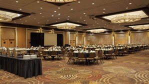 Une salle de banquet avec des tables rondes dressées pour un événement formel, dotée d’un tapis à motifs et d’un plafond haut orné de lustres.