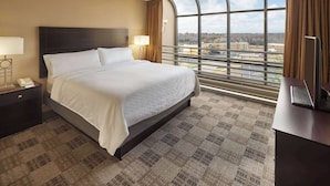 Une chambre d’hôtel dotée d’un grand lit, de deux tables de chevet avec des lampes, d’un bureau avec un ordinateur et d’une grande fenêtre donnant sur la ville.