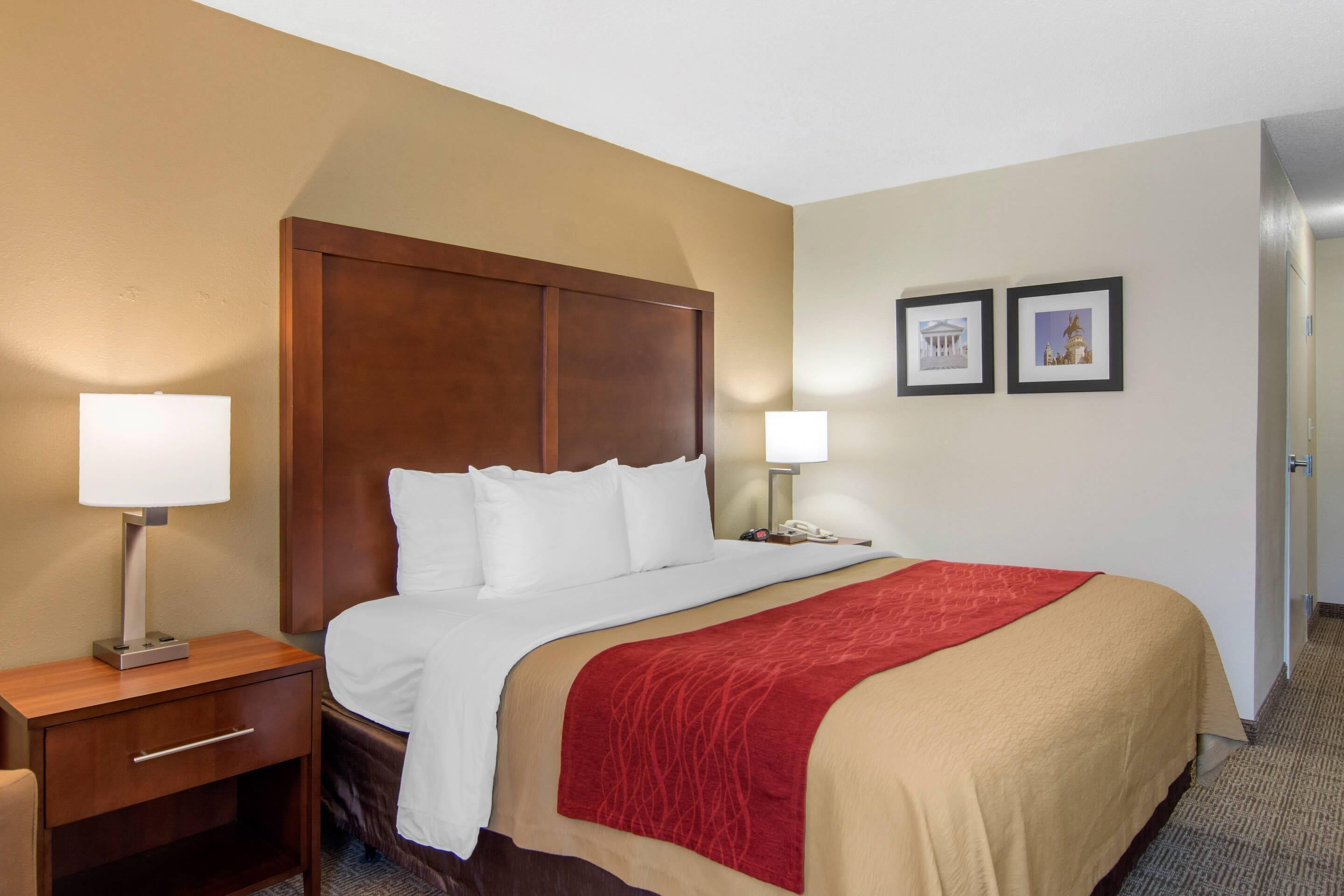 A hotel room with a bed, bedside tables, a red throw blanket, and two framed pictures on the wall.