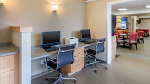 A modern hotel room with a desk, two monitors, a keyboard, and a chair. There is a dining area with tables and chairs in the background. - Comfort Inn Chester - Richmond South (Chester)