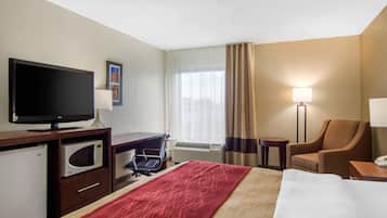 Ein Hotelzimmer mit einem Bett, einem Schreibtisch mit Fernseher, einer Mikrowelle, einem Stuhl, einer Lampe und einem Fenster mit Vorhängen.