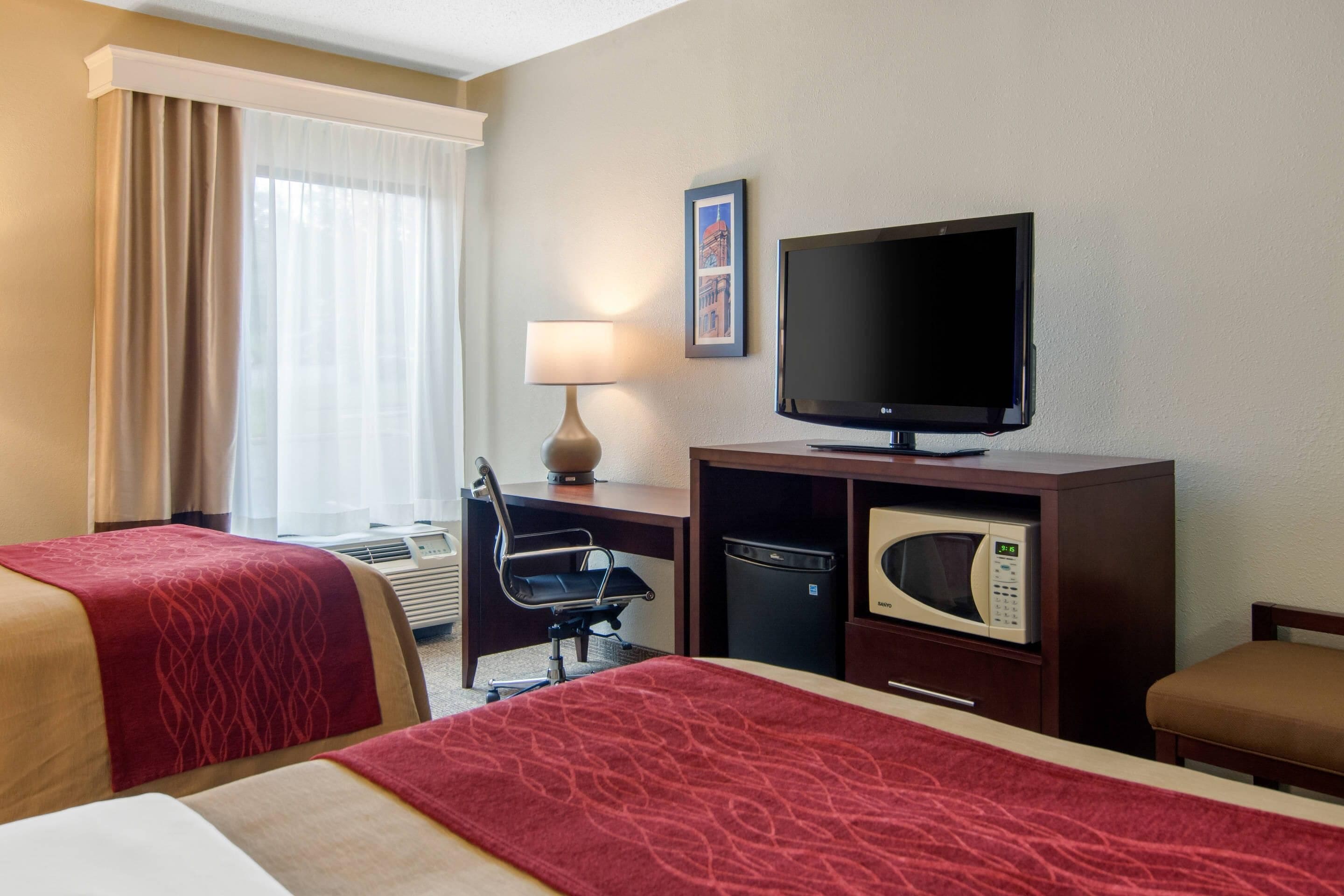 A hotel room with two beds, a desk, a television, a microwave, and a window with curtains.