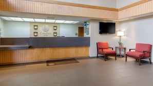 A hotel reception area with a wooden reception desk, a flat-screen TV, and two red chairs.