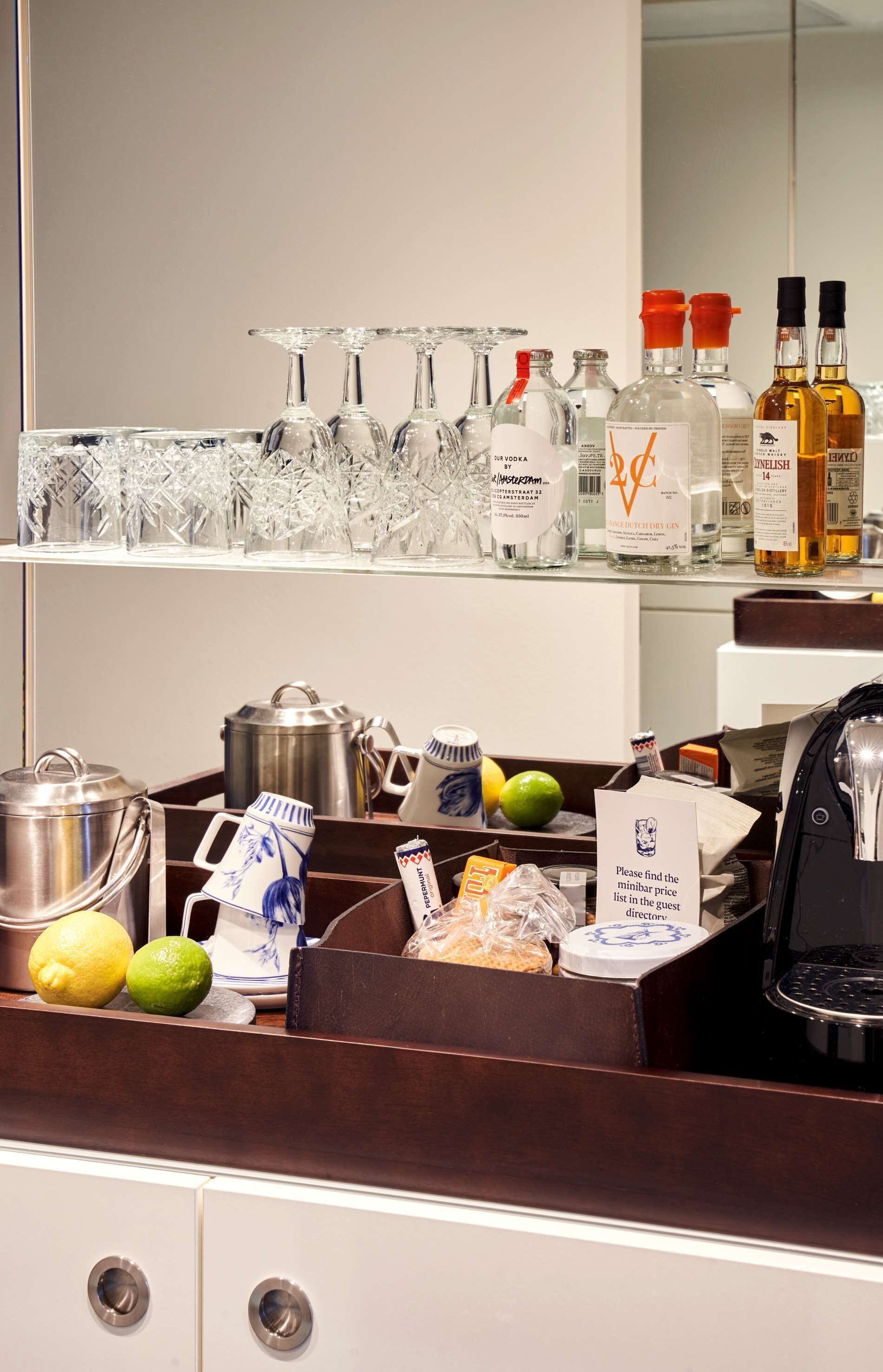 A hotel minibar with various beverages, glasses, and a sign instructing to find the minibar price list in the guest room.