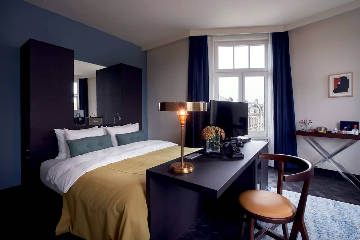 A hotel room with a bed, a desk, a chair, a lamp, a window with curtains, and a view of a building outside.