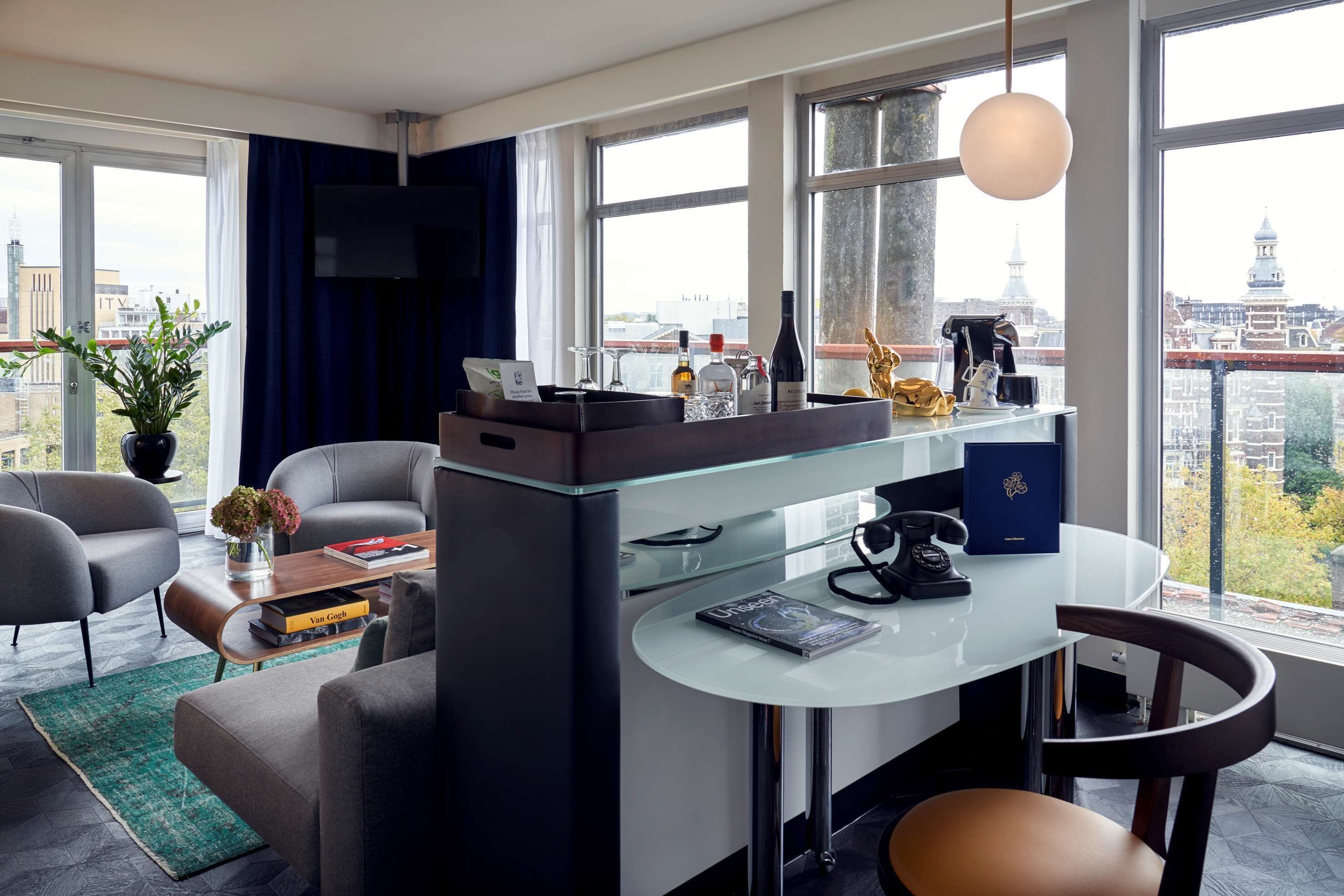 A modern hotel room with a central island, seating area, and large windows offering city views.