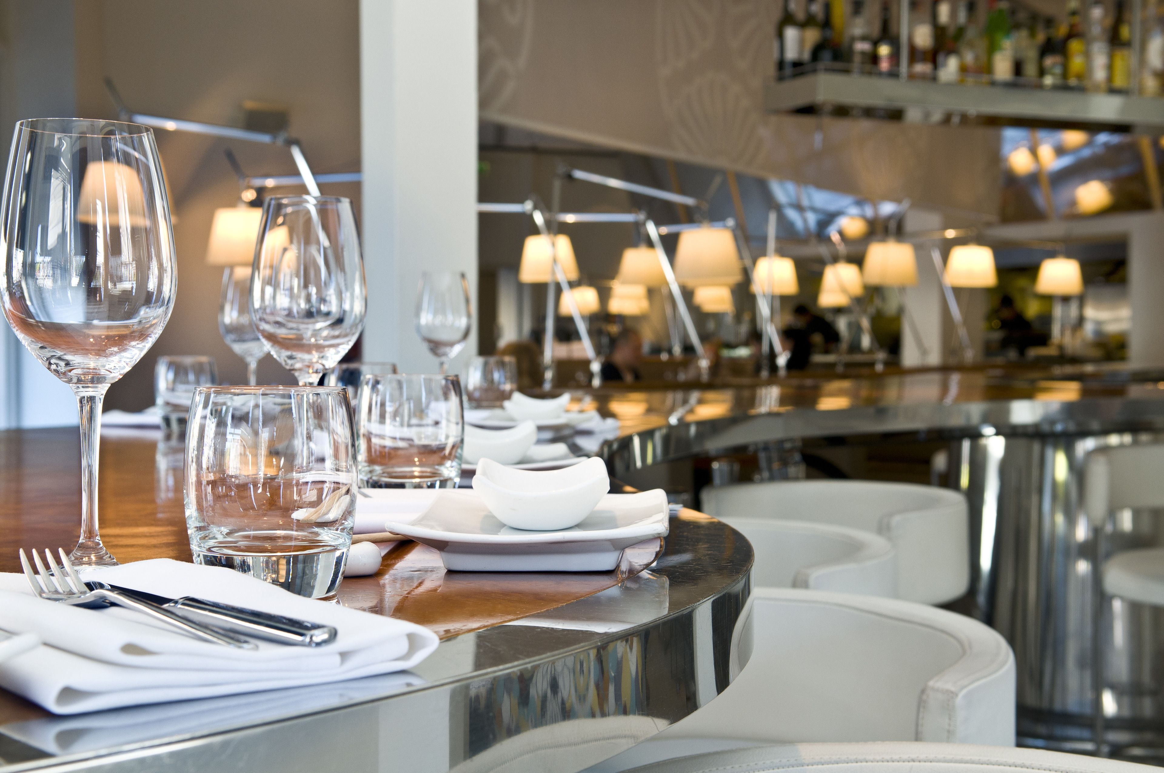 A restaurant setting with a bar area, white chairs, and a well-lit ambiance with hanging lamps.