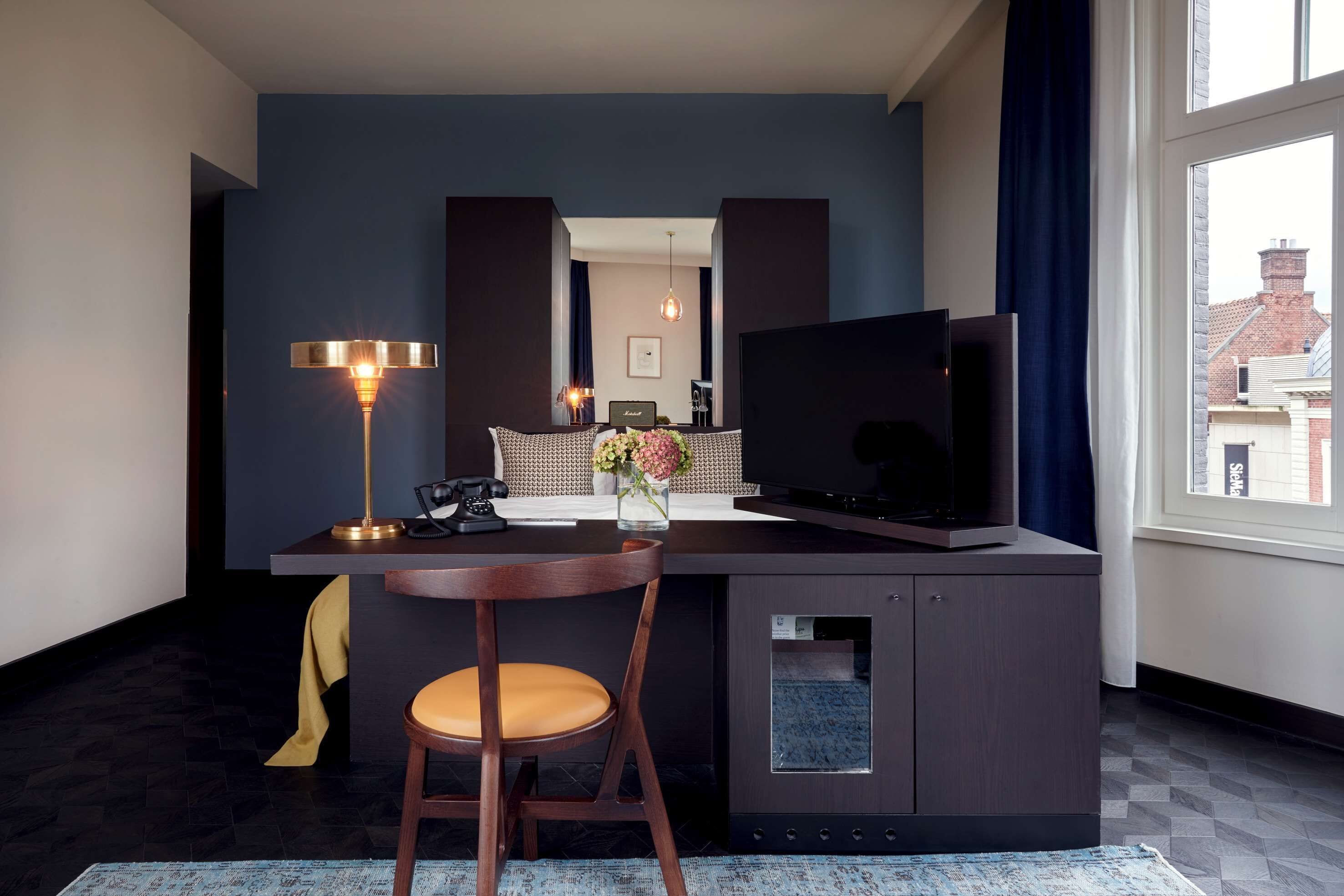 A modern hotel room with a dark blue accent wall, a large window with curtains, a flat-screen TV, a desk with a chair, a lamp, and a small cabinet.
