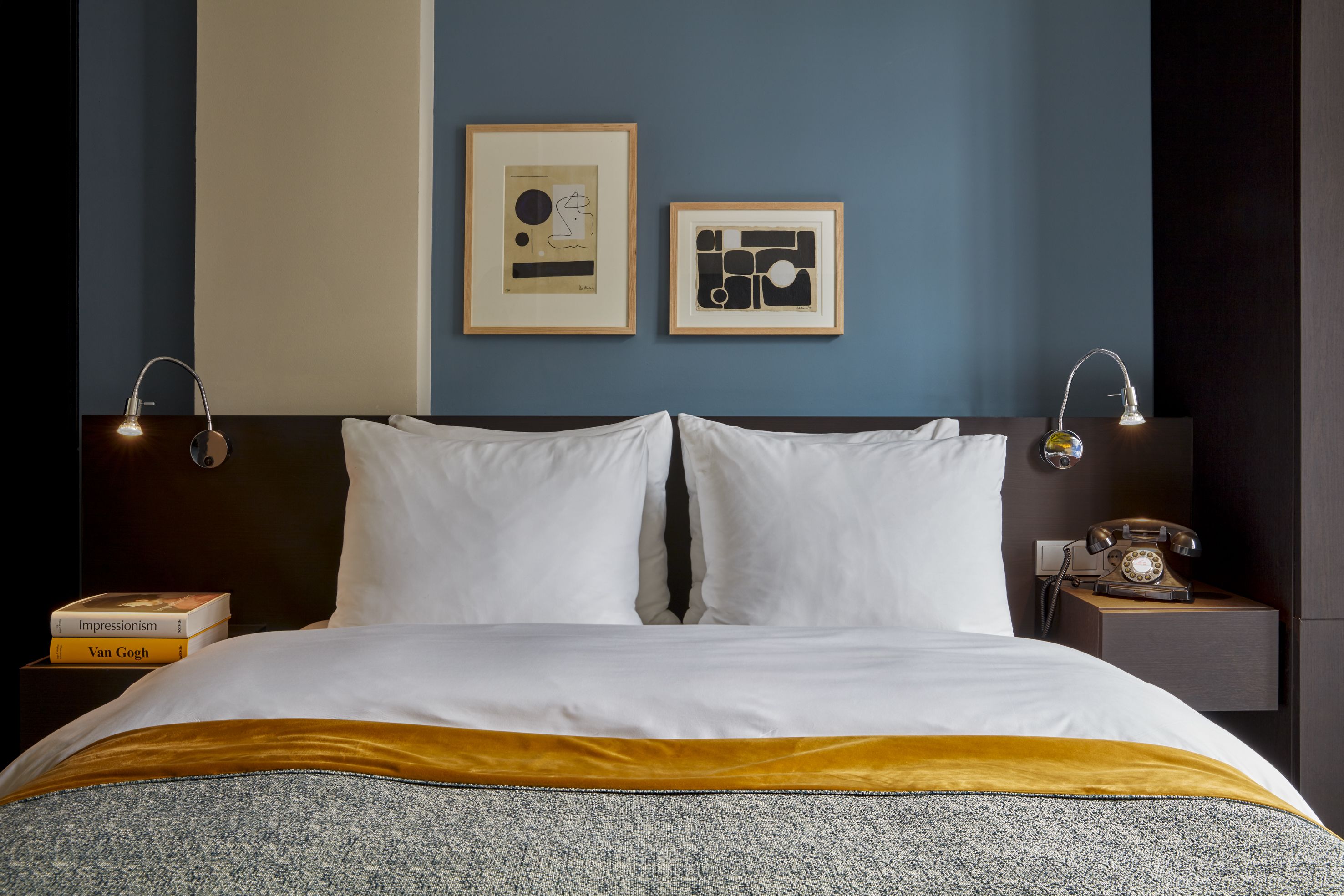 Een net opgemaakt bed met wit en goud linnengoed, twee ingelijste abstracte kunstwerken aan de muur en een vintage draaischijf telefoon op het nachtkastje.