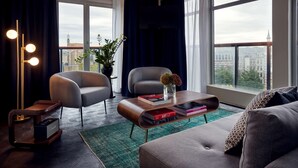 Ein modernes Wohnzimmer mit einer Couch, zwei Sesseln, einem Couchtisch und einer Stehlampe. Durch große Fenster ist ein Blick auf Gebäude möglich.