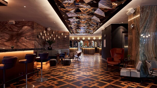 Eine moderne Lobby mit einem geometrischen Fußbodenmuster, einem Empfangsbereich mit Marmorwand und verschiedenen Sitzgelegenheiten, darunter Barhocker und Sessel.
