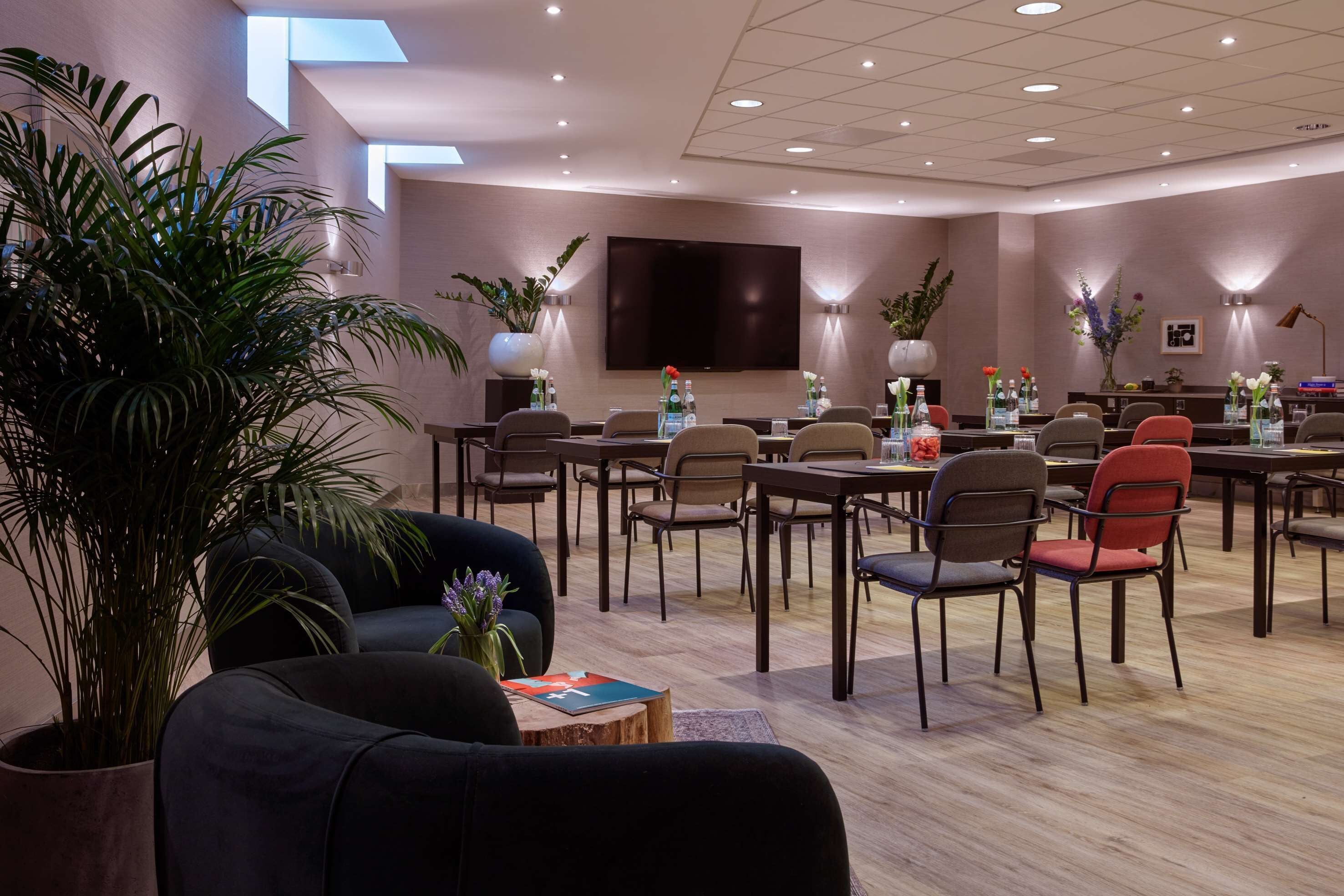A modern conference room with a large flat-screen TV, multiple tables with chairs, and potted plants.