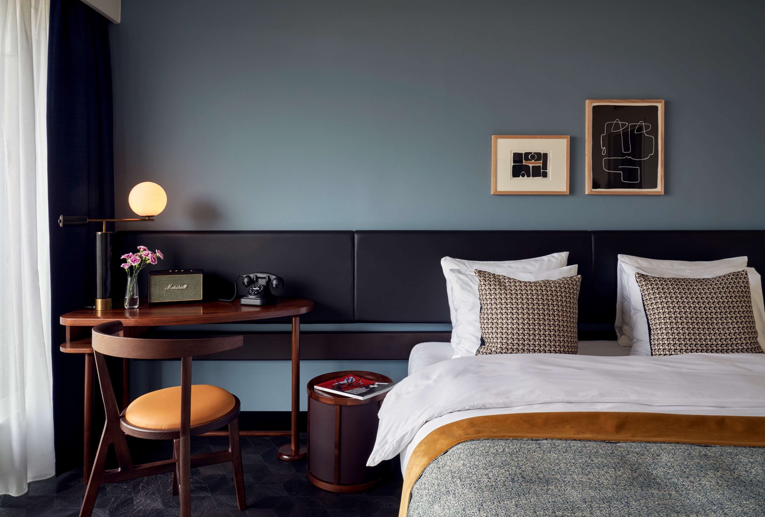A hotel room with a bed, a desk with a lamp, a chair, and two framed pictures on the wall.