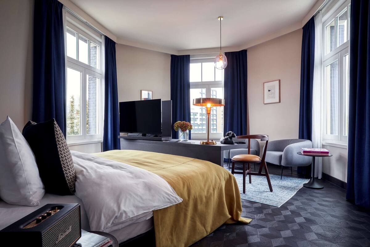 A hotel room with a bed, a desk, a television, a chair, and large windows with curtains.