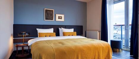 Habitación de hotel con una cama grande, una mesita de noche, una lámpara y vistas de la ciudad a través de un ventanal.
