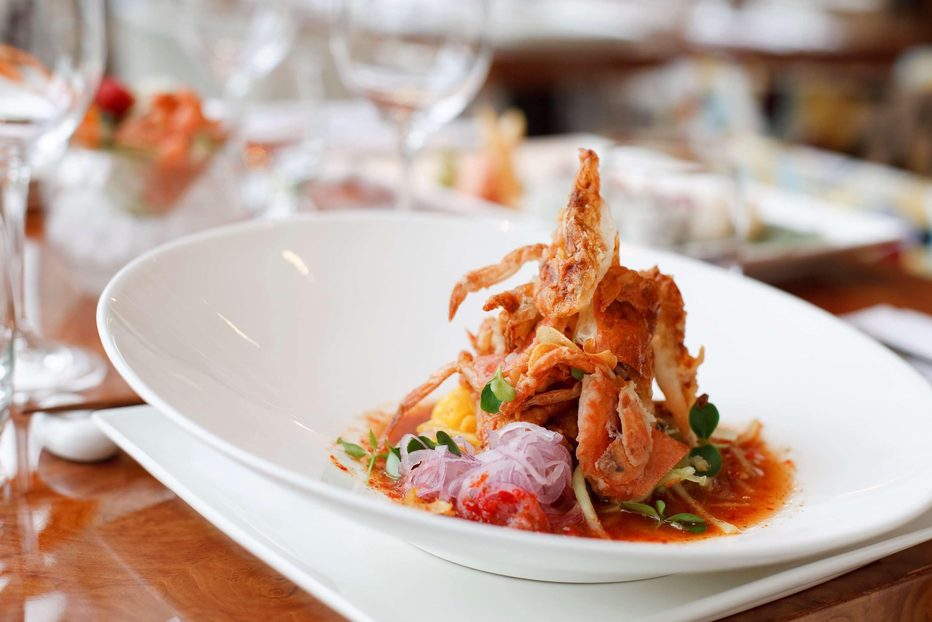 Un plato blanco con un plato de mariscos, posiblemente cangrejo, adornado con brotes diminutos y salsa roja, sobre una mesa de madera.