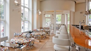Eine moderne Bar mit einer langen Theke und weißen Barhocker. Bunte, gemusterte Ottomane und ein großes Fenster sorgen für natürliches Licht.