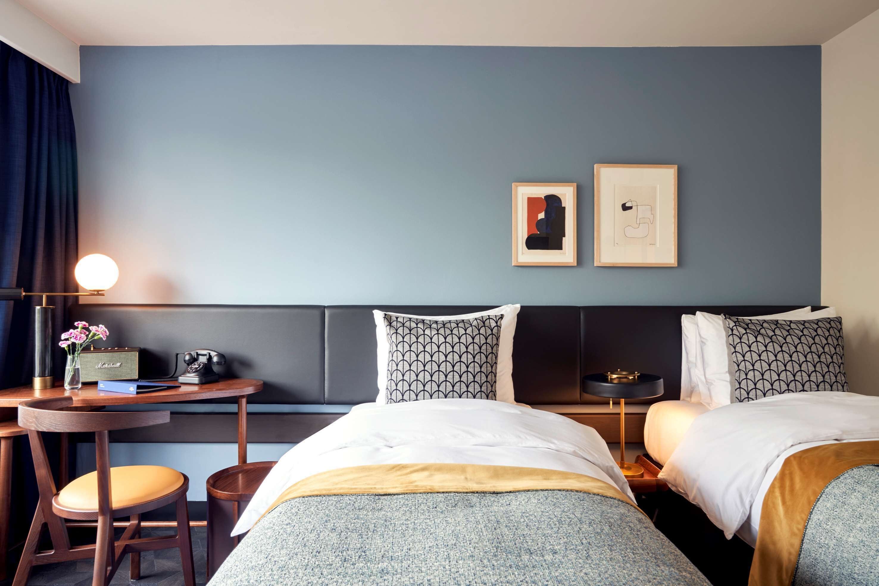 Habitación de hotel con dos camas, un escritorio con lámpara, una silla y una mesita con radio y flores.