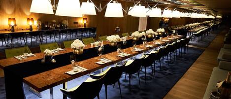 Una larga mesa de comedor con sillas, adornada con flores blancas y copas de vino, en una sala bien iluminada con candelabros modernos.
