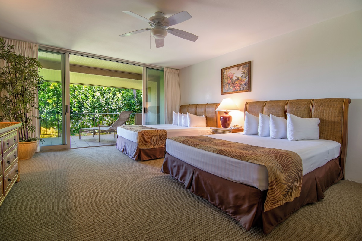 Habitación de hotel con dos camas, ventilador de techo, balcón con vistas a la vegetación y cómoda de madera.