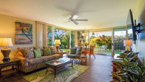 A spacious living room with a sofa, coffee table, and chairs, a ceiling fan, and large windows offering a view of the outdoors.