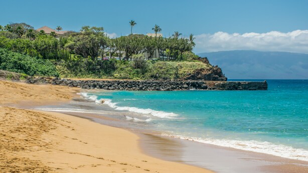 A sandy beach with clear turquoise water, a rocky breakwater, and lush greenery in the background.
